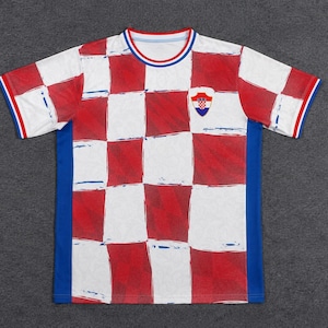 Może przedstawiać: Koszulka z kr&oacute;tkim rękawem i okrągłym dekoltem, ozdobiona czerwono-białym wzorem w szachownicę z przodu. T-shirt posiada jednolite niebieskie panele boczne, niebiesko-czerwony prążkowany kołnierzyk oraz pasujące mankiety z małym herbem Chorwacji.