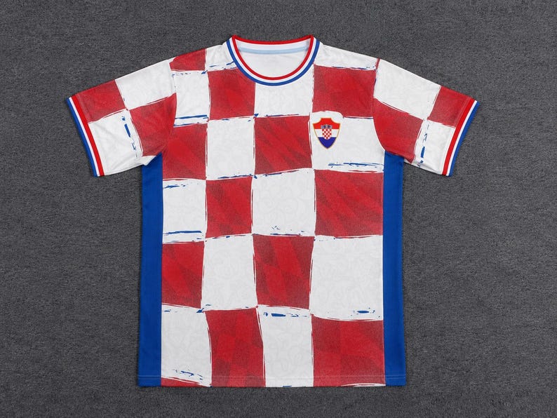 Może przedstawiać: Koszulka z kr&oacute;tkim rękawem i okrągłym dekoltem, ozdobiona czerwono-białym wzorem w szachownicę z przodu. T-shirt posiada jednolite niebieskie panele boczne, niebiesko-czerwony prążkowany kołnierzyk oraz pasujące mankiety z małym herbem Chorwacji.