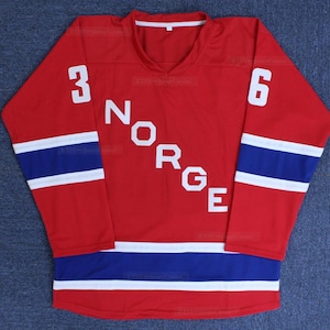 以下が含まれることがあります： 白文字で「NORGE」と数字の「3」と「6」が入った赤いホッケーのジャージ。袖と裾に青と白のストライプが入っています。通気性の良い素材で作られています。