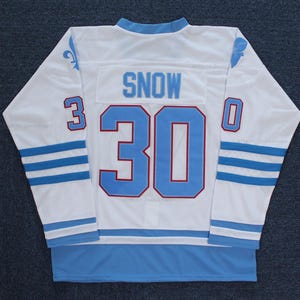 Könnte beinhalten: Ein weißes Hockeytrikot mit blauen und roten Akzenten. Die Nummer 30 ist auf der Rückseite mit dem Namen "SNOW" darüber. Das Trikot hat einen blauen Schmetterling auf der rechten Schulter und eine Lilie auf der linken Schulter.