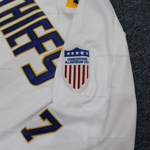 May include: White hockey jersey with blue and gold lettering that reads "Chief's" and the number "7". The jersey has a patch on the sleeve that reads "Charlestown All-American City" with a shield and five stars above the text and a red, white, and blue American flag inside the shield.