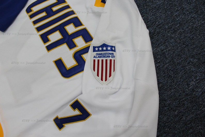 May include: White hockey jersey with blue and gold lettering that reads "Chief's" and the number "7". The jersey has a patch on the sleeve that reads "Charlestown All-American City" with a shield and five stars above the text and a red, white, and blue American flag inside the shield.