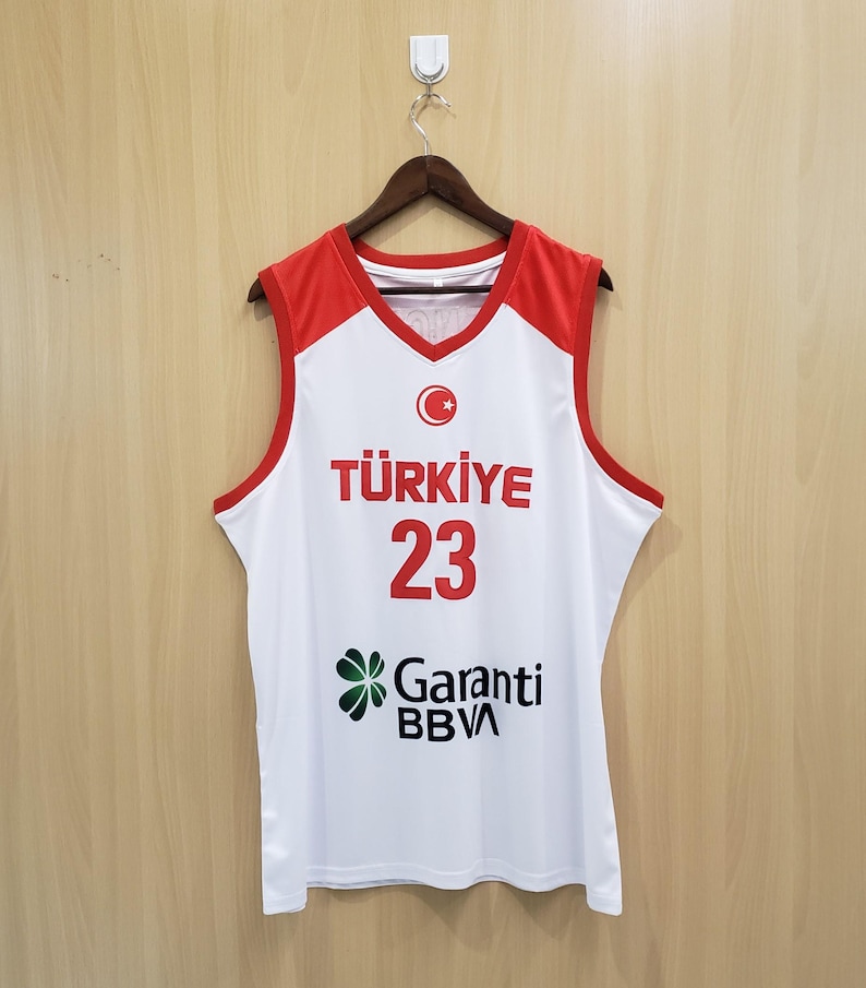 K&ouml;nnte beinhalten: Ein wei&szlig;es und rotes Basketballtrikot mit dem Wort "T&Uuml;RKIYE" in Rot, der Nummer "23" und dem Logo "Garanti BBVA". Das Trikot hat einen V-Ausschnitt und ist &auml;rmellos.