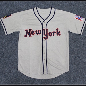 May include: A gray baseball jersey with the word "New York" in red and black lettering on the front. The jersey has navy blue trim and a button-up front. There are patches on the sleeves.