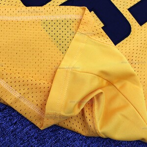May include: Close-up of the yellow mesh fabric of a jersey with a navy blue number sewn on. The fabric is folded over and the stitching is visible.