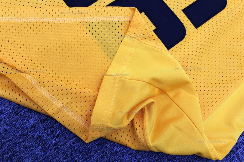 May include: Close-up of the yellow mesh fabric of a jersey with a navy blue number sewn on. The fabric is folded over and the stitching is visible.