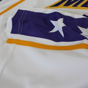 May include: Close-up of a white jersey with a purple and gold stripe. The stripe features two white stars.