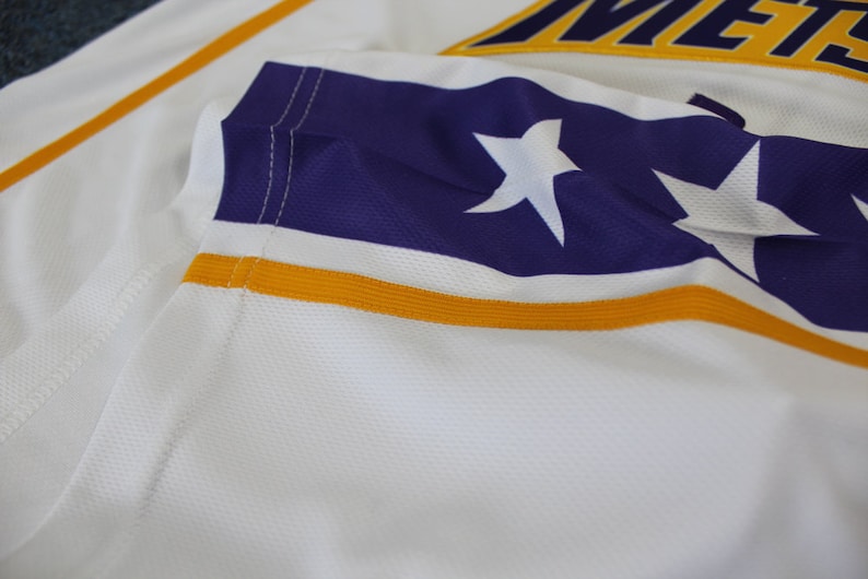 May include: Close-up of a white jersey with a purple and gold stripe. The stripe features two white stars.