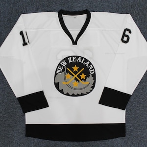 May include: White hockey jersey with black accents, featuring the text "NEW ZEALAND" around a logo with stars and crossed hockey sticks. The jersey has the number "1" on the left sleeve and "6" on the right sleeve.