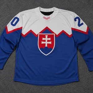 Könnte beinhalten: Ein langärmeliges Eishockeytrikot mit weißem Oberkörper und Schulterbereich sowie einem blauen Unterteil. Die Zahl 02 ist auf den Schultern aufgedruckt. Ein rot-weiß-blaues Wappen mit einem Doppelkreuz ist mittig auf der Brust platziert.