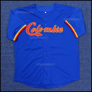 Pode incluir: Uma camisa de beisebol azul com a palavra "Colômbia" em escrita vermelha no peito. As mangas têm listras amarelas e vermelhas. A camisa tem uma frente abotoada.
