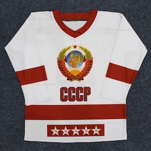 Peut inclure: Un maillot de hockey blanc avec des rayures rouges et blanches. Le maillot présente un grand emblème brodé des armoiries de l'Union soviétique avec une faucille et un marteau, un globe terrestre et des épis de blé. Le texte "CCCP" est imprimé en noir sous l'emblème. Cinq étoiles rouges avec une faucille et un marteau sont imprimées en bas du maillot.