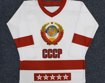 May include: White hockey jersey with red and white stripes. The jersey features a large embroidered emblem of the Soviet Union coat of arms with a hammer and sickle, a globe, and wheat stalks. The text "CCCP" is printed in black below the emblem. Five red stars with a hammer and sickle are printed on the bottom of the jersey.