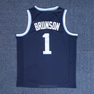 May include: Navy blue basketball jersey with white lettering and the name "BRUNSON" above the number "1". The jersey has light blue trim around the collar and arm openings. The jersey is displayed on a gray surface.