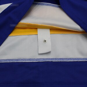 May include: Close-up of the inside of a blue and yellow hockey jersey. The jersey has a white strip with a white button and a snap closure.