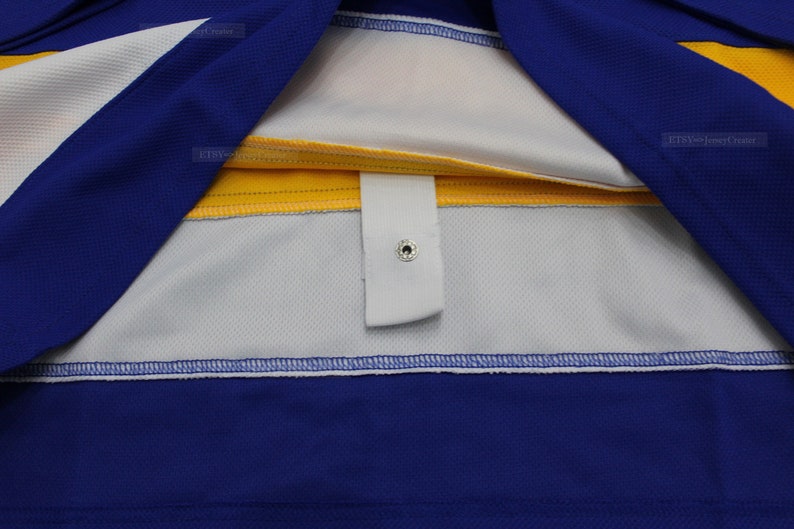 May include: Close-up of the inside of a blue and yellow hockey jersey. The jersey has a white strip with a white button and a snap closure.