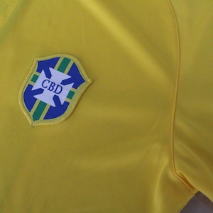 May include: Close-up of a yellow shirt with a shield-shaped emblem. The emblem features a blue and green border with a white cross design and the letters "CBD" in white. The shirt is made of a smooth fabric.