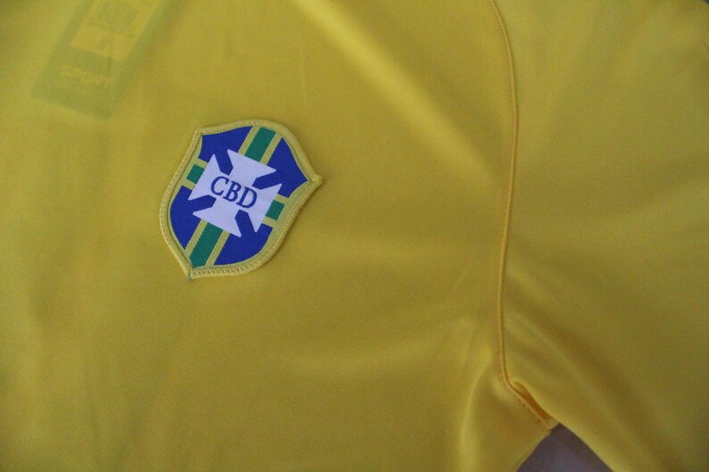 May include: Close-up of a yellow shirt with a shield-shaped emblem. The emblem features a blue and green border with a white cross design and the letters "CBD" in white. The shirt is made of a smooth fabric.