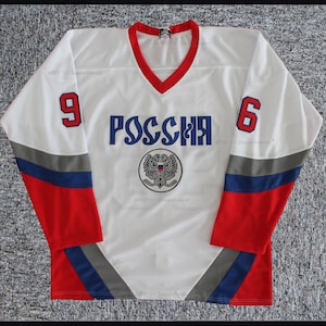 Könnte beinhalten: Ein weißes Hockeytrikot mit dem Wort "Россия" (Russland) in blauer Schrift und dem russischen Wappen in der Mitte. Das Trikot hat rote, blaue und graue Streifen an den Ärmeln und die Nummern "9" und "6" auf den Schultern.