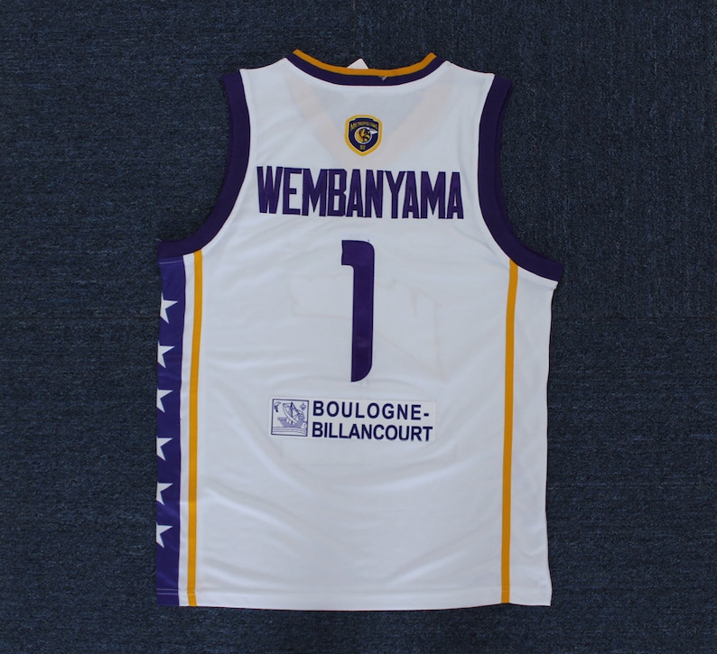 May include: White basketball jersey with purple and gold trim. The back of the jersey has the number 1 and the name "Wembanyama" printed in purple. The team name "BOULOGNE-BILLANCOURT" is printed in a small box on the bottom of the jersey.