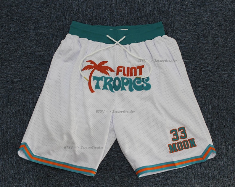 Puede incluir: Pantalones cortos de baloncesto de malla blanca con cintura verde y ribetes naranjas y turquesas. Los pantalones cortos presentan el texto "FLINT TROPICS" con un gr&aacute;fico de palmera y el n&uacute;mero "33 MOON" en naranja.