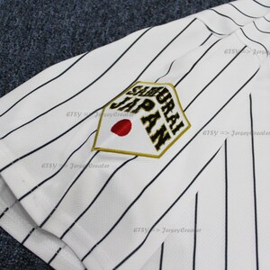 May include: White baseball jersey with black pinstripes and a gold and black embroidered patch that says "Samurai Japan" with a red circle in the center. The jersey also has a large black and gold "J" on the front.