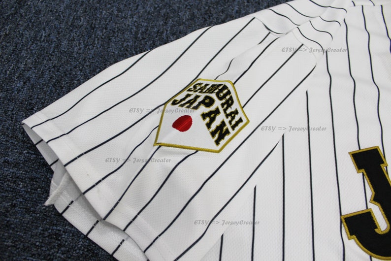 May include: White baseball jersey with black pinstripes and a gold and black embroidered patch that says "Samurai Japan" with a red circle in the center. The jersey also has a large black and gold "J" on the front.