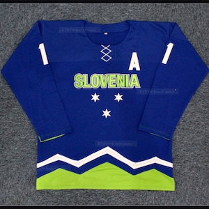 Könnte beinhalten: Ein blaues Langarm-Trikot mit dem Wort "SLOVENIA" in Grün und Weiß sowie drei weißen Sternen. Das Trikot hat weiße Zahlen auf den Ärmeln und einen weißen Schnürkragen. Der untere Teil hat ein grün-weißes Design.