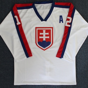Könnte beinhalten: Weißes Hockeytrikot mit roten und blauen Besätzen. Das Trikot zeigt das slowakische Staatswappen mit einem roten Kreuz und blauen Wolken auf einem weißen Schild. Die Nummern "1" und "A2" befinden sich auf den Ärmeln.