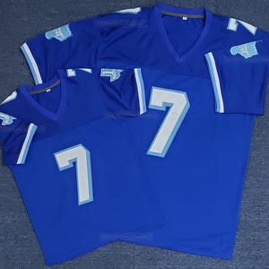 May include: Three blue football jerseys with white and light blue accents. Each jersey has the number 7 on the front and a patch on the sleeve. The jerseys have a V-neck design.