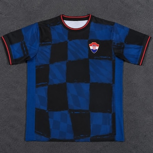 May include: A short-sleeve t-shirt featuring a dark blue and black checkered pattern across the front and sleeves. The crew neck collar and sleeve cuffs are trimmed with a thin red line. A small shield-shaped emblem with a red and white checkerboard design is positioned on the upper left chest area.