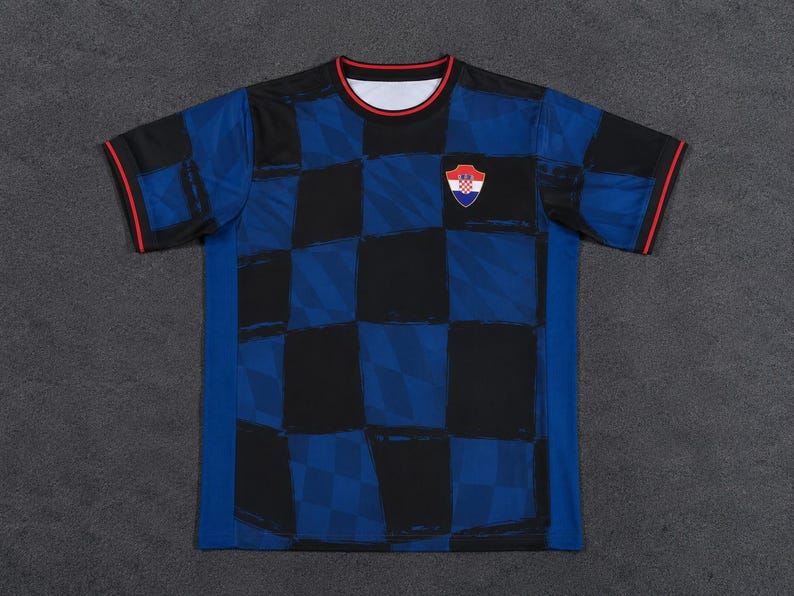 May include: A short-sleeve t-shirt featuring a dark blue and black checkered pattern across the front and sleeves. The crew neck collar and sleeve cuffs are trimmed with a thin red line. A small shield-shaped emblem with a red and white checkerboard design is positioned on the upper left chest area.