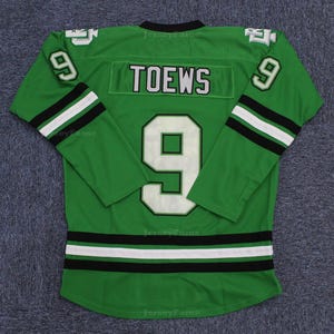 Puede incluir: Jersey de hockey verde con el número 9 blanco en la parte delantera, trasera y mangas. El nombre "TOEWS" se muestra en la parte superior de la espalda. Rayas blancas y negras acentúan las mangas y el dobladillo inferior. El jersey tiene un cuello negro.