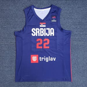 May include: A sleeveless navy blue basketball jersey with a V-neck. The jersey features the word "SRBIJA" in white block letters, the number "22" in red, and the "triglav" logo. A Serbian flag is above the word "SRBIJA".