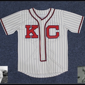 May include: White baseball jersey with blue and white stripes. The jersey has red lettering that says "KC".