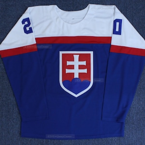 Könnte beinhalten: Ein blaues und weißes Hockeytrikot mit dem slowakischen Staatswappen auf der Vorderseite. Das Trikot hat rote Streifen an den Ärmeln und einen weißen Kragen.