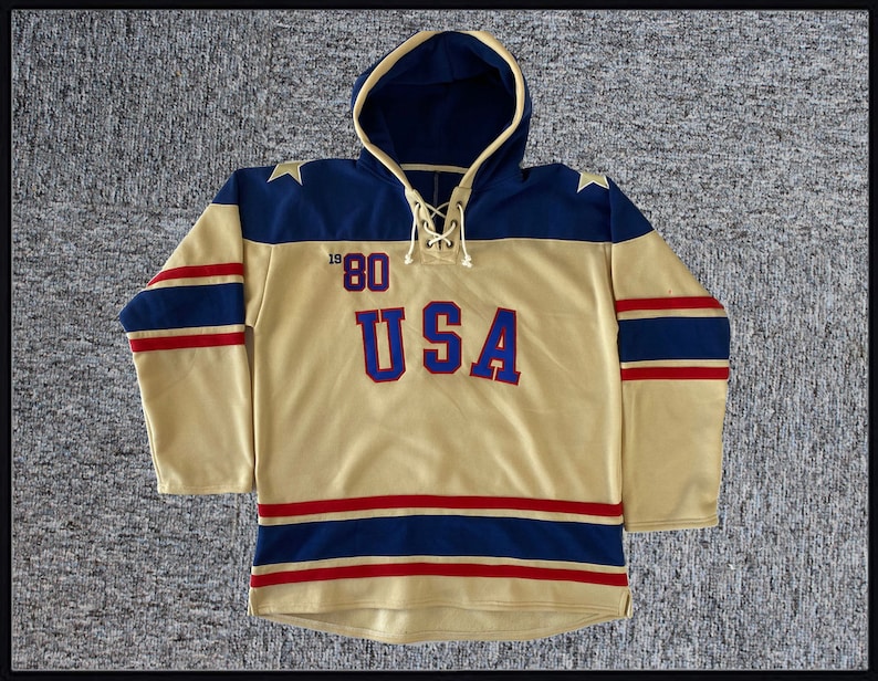 May include: A tan hooded sweatshirt with blue and red stripes. The sweatshirt has a large "USA" graphic on the front with the number "80" above it.