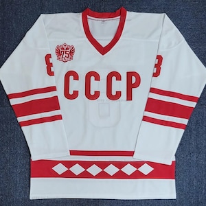 May include: White hockey jersey with red accents, featuring the letters "CCCP" in large red block letters across the chest. The jersey has red stripes on the sleeves and a red V-neck collar. A crest with the number "75" is on the chest.