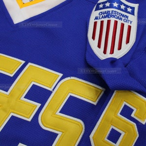 May include: A blue hockey jersey with a yellow number "56" on the front. The jersey has a patch on the right shoulder that says "Charlestown All-American City" with a red, white, and blue American flag design.