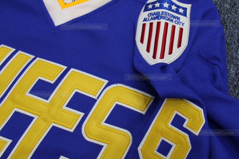 May include: A blue hockey jersey with a yellow number "56" on the front. The jersey has a patch on the right shoulder that says "Charlestown All-American City" with a red, white, and blue American flag design.