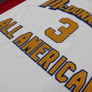 May include: White jersey with gold and blue lettering. The words "McDonald's" and "All American" are visible, along with the number "3". The jersey has a red stripe along the top.