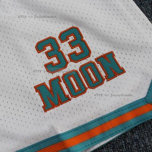Puede incluir: Pantalones cortos deportivos de malla blanca con detalles en turquesa y naranja. Los pantalones cortos presentan el n&uacute;mero "33" y el nombre "MOON" en turquesa con ribete naranja. El ribete de los pantalones cortos es turquesa y naranja. El nombre de la tienda Etsy "JerseyCreator" es visible.