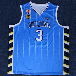 May include: A light blue basketball jersey with the word "BEIJING" and the number "3" in white. The jersey has a V-neck and white trim. Vertical white stripes run down the front. Logos for CBA and UPS are visible.