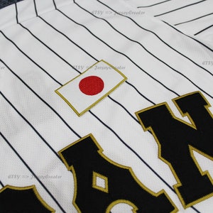 May include: White baseball jersey with black pinstripes and a large black "JAPAN" lettering with gold trim. The jersey has a small Japanese flag patch on the chest.