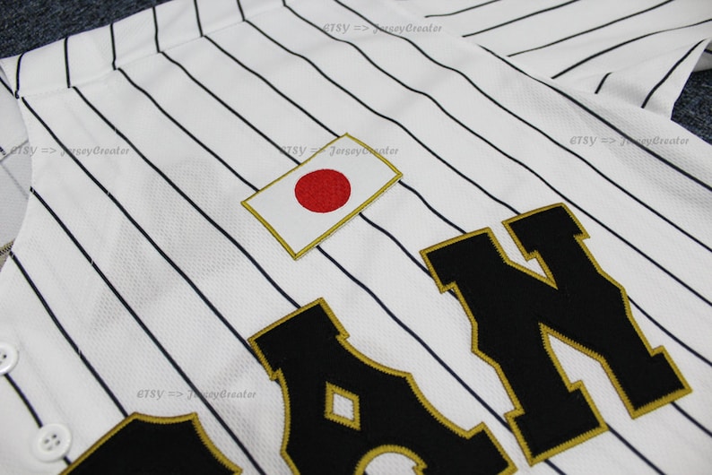 May include: White baseball jersey with black pinstripes and a large black "JAPAN" lettering with gold trim. The jersey has a small Japanese flag patch on the chest.