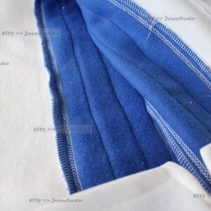 K&ouml;nnte beinhalten: Nahaufnahme des Innenfutters eines wei&szlig;en und blauen Kapuzenpullovers. Das blaue Futter ist sichtbar und zeigt die N&auml;hte und das weiche Fleece-Material.