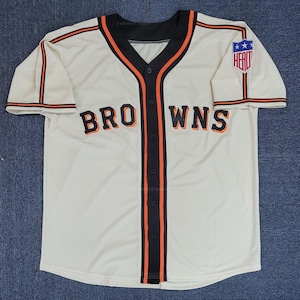 Puede incluir: Camiseta de béisbol color crema con la palabra "BROWNS" en el pecho en letras negras con ribete naranja. La camiseta tiene ribetes negros y naranjas alrededor del cuello, en la parte delantera y en las mangas. Un parche con la palabra "HEAT" está en la manga derecha.