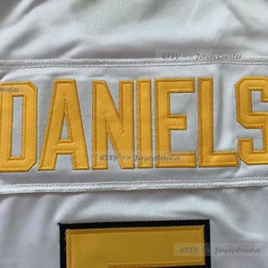 Op de afbeelding: Wit shirt met een gele naamplaat met de tekst "DANIELS".