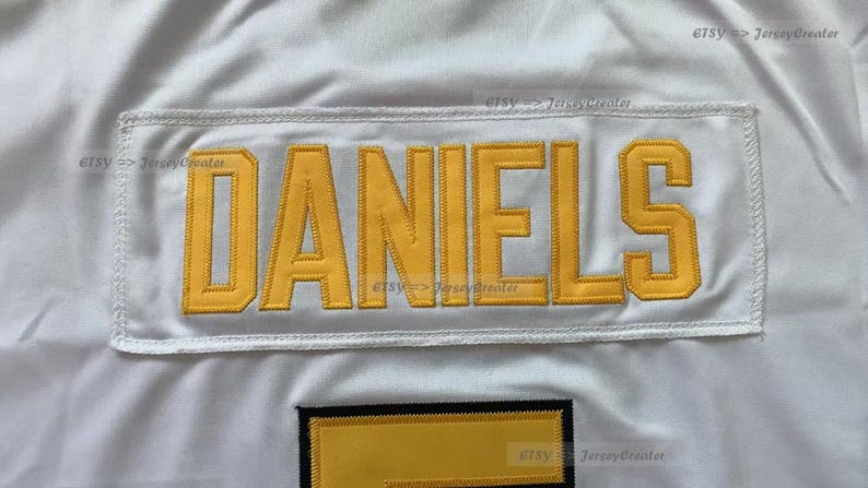 Op de afbeelding: Wit shirt met een gele naamplaat met de tekst "DANIELS".
