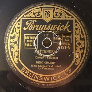 Puede incluir: Un disco de vinilo negro con una etiqueta dorada. La etiqueta presenta el logotipo de Brunswick, el título de la canción "Jattendrai", el artista Bing Crosby y el número de disco 0121-A.